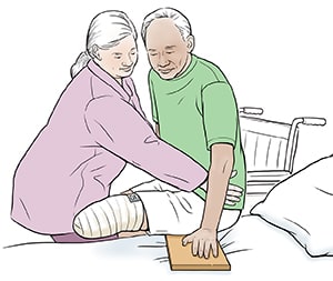 Health care provider helping man with amputated leg to use slide board from wheelchair into bed.