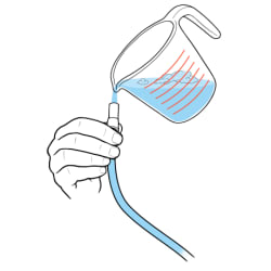 Hand holding opening of catheter bag tube with measuring cup pouring soapy water into tube.