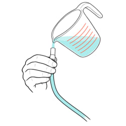 Hand holding opening of catheter bag tube with measuring cup pouring cleaning solution into tube.